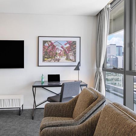Meriton Suites Herschel Street, Brisbane Exterior foto The photo shows a modern room interior that includes two comfortable chairs with a textured fabric, a television mounted on the wall, and a desk featuring a laptop and a lamp. A piece of artwork is displayed on the wall above the desk. Large windows 