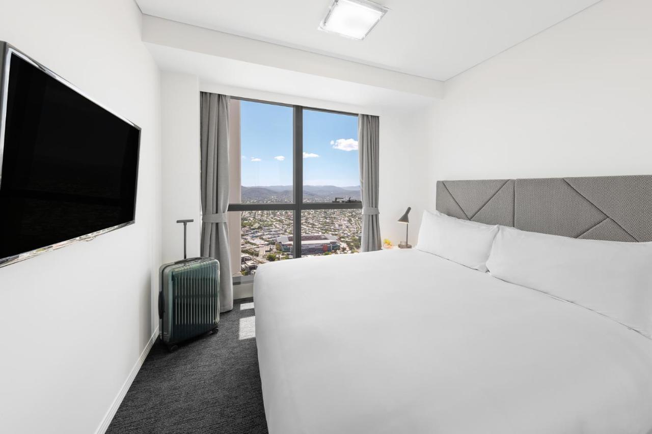 Meriton Suites Herschel Street, Brisbane Exterior foto The photo shows a modern hotel room. It features a large bed with white bedding and a gray headboard. There's a flat-screen TV mounted on the wall across from the bed. To the side, there's a small luggage trolley with a suitcase. The room has large w